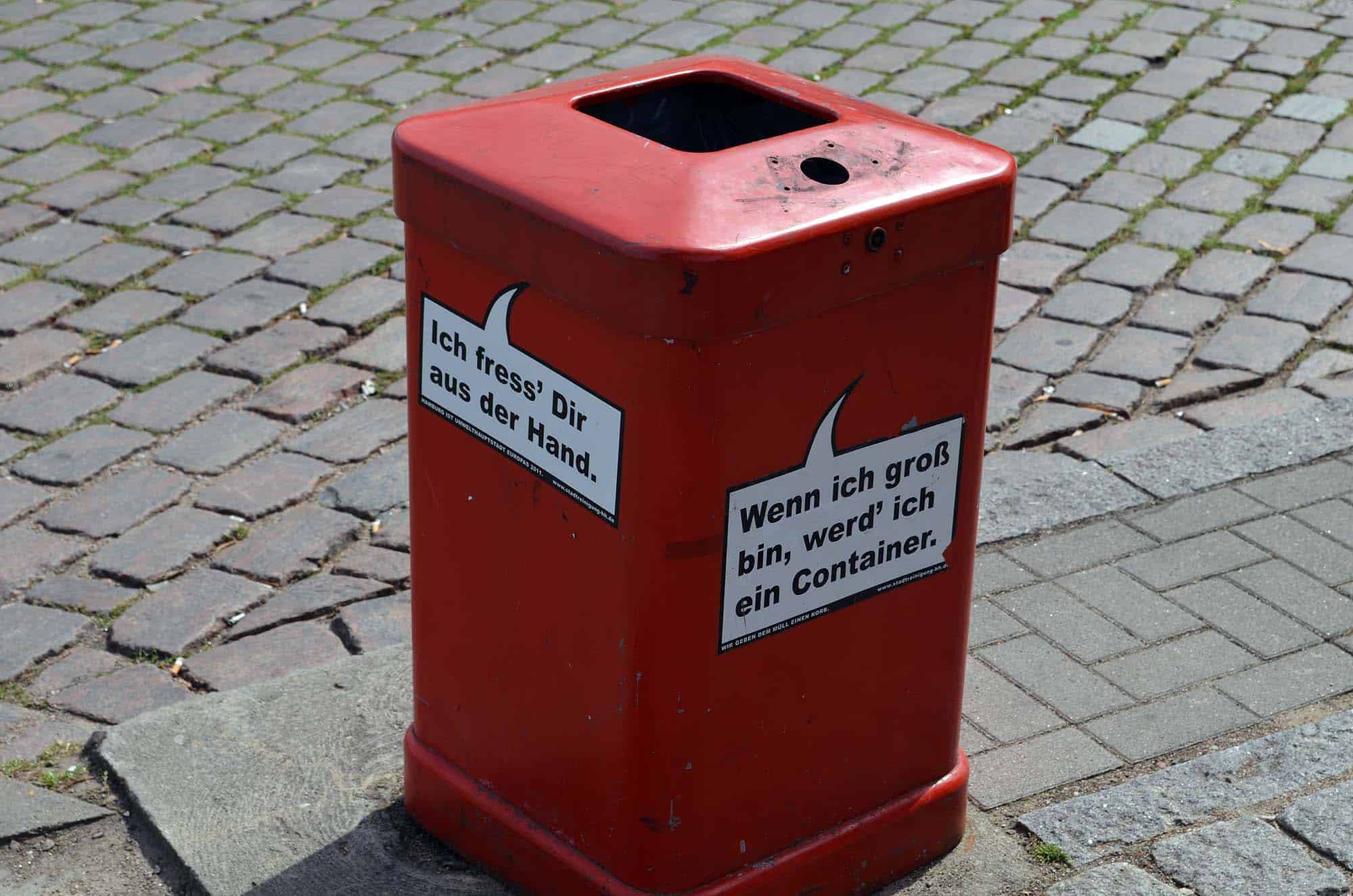 Roter Mülleimer mit witzigen Sprüchen auf dem Pflasterweg.